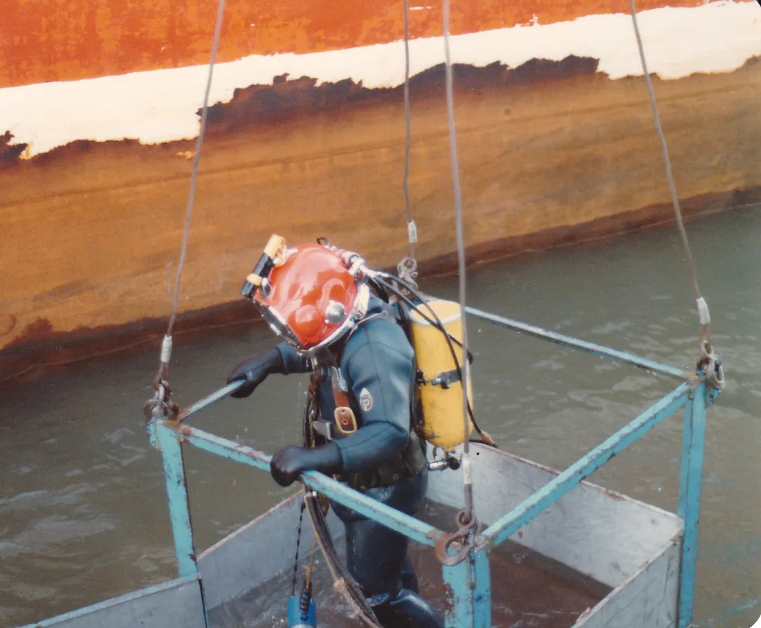 A diver in a red helmet riding the dive stage beside a ship's hull